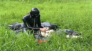 Fetish slave in latex walks and sucks a dildo on the grass